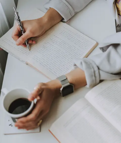 A person writing in a notebook while holding a coffee mug